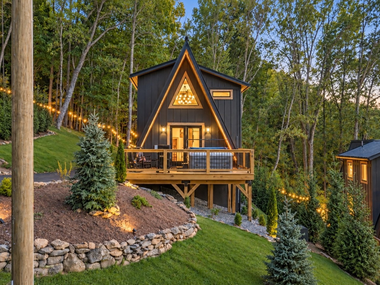 The West Jefferson A-frame cabin at dusk with string lights and a stone-edged lawn.