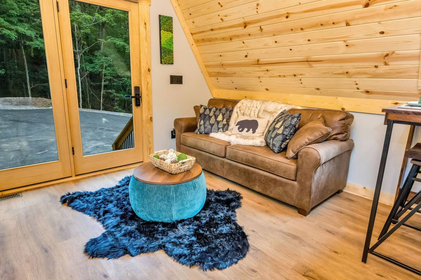 Living nook with leather loveseat, teal ottoman, and glass doors to the woods